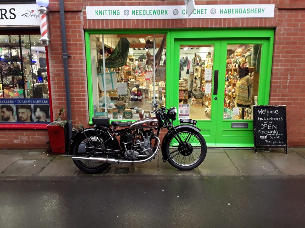 Shop front of Pins & Needles (Devizes). Modern brick build show with green woodwork around doors and window. Large window and double doors. Pavement is one and a bit flagstones wide.